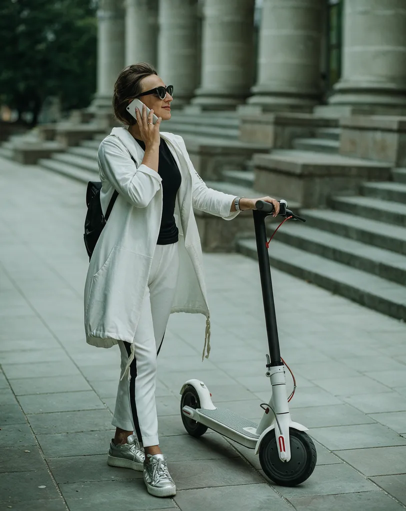 Femme au téléphone tenant sa trottinette électrique blanche devant un bâtiment